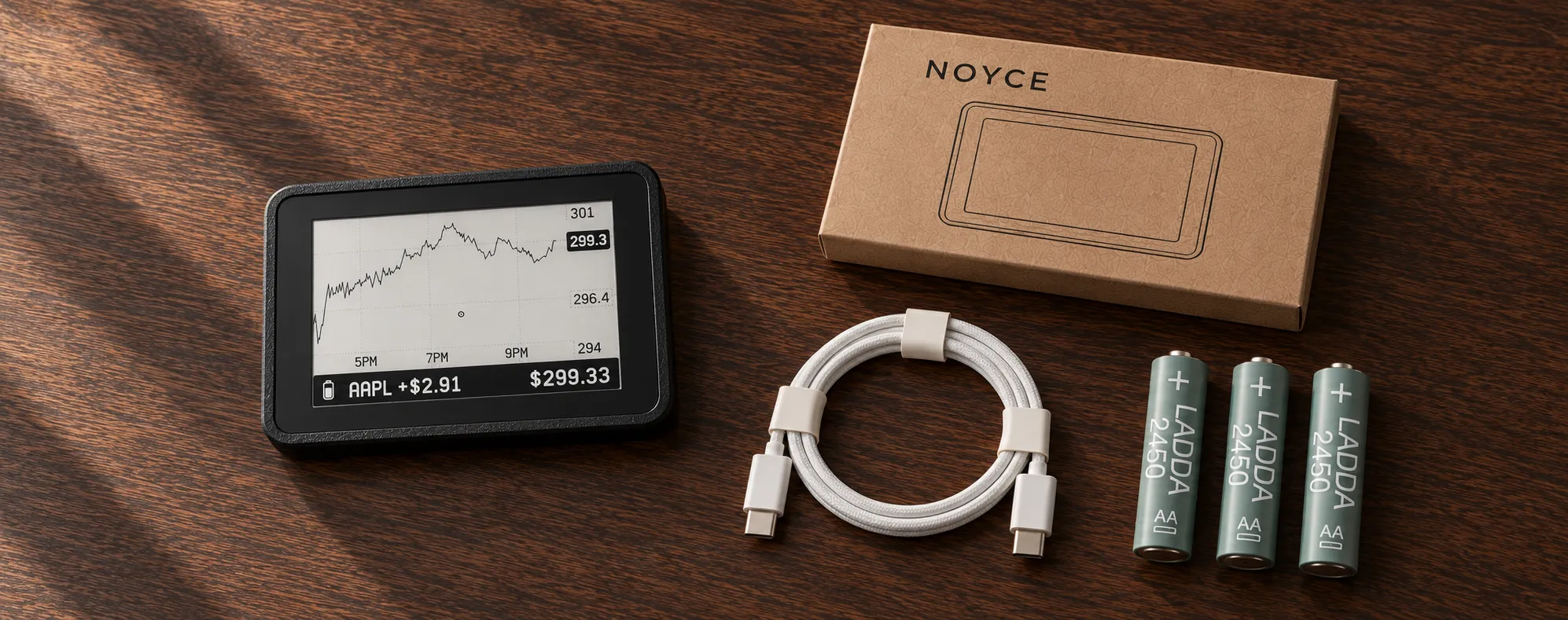 NOYCE One e-paper display, kraft paper box, white braided USB-C cable and three IKEA LADDA rechargeable AA batteries arranged on a wooden table